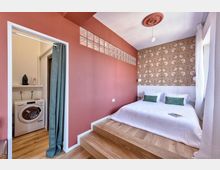 Camera da letto moderna con letto su pedana rialzata in legno, parete color terracotta con fascia di mattoni di vetro e testata con carta da parati floreale. A sinistra si apre una nicchia lavanderia con lavatrice e tenda, mentre sulla destra una finestra con tende bianche illumina l’ambiente.