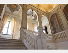 Ampio scalone interno in uno stile classico con balaustre in marmo bianco, pareti ornate da archi decorati e soffitti affrescati. Le finestre sono adornate con tende chiare che filtrano la luce naturale.