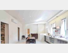 Cucina moderna e luminosa con pareti bianche, soffitto alto con faretti e grandi finestre sul lato destro. Lungo la parete destra ci sono mobili e elettrodomestici in acciaio con piano di lavoro, mentre al centro si trova un tavolo da pranzo in legno con sedie e un piccolo vaso di fiori.