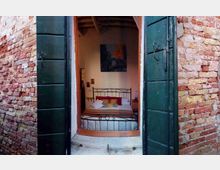 Finestra aperta con scuri verdi su una parete in mattoni a vista, che affaccia su una camera da letto interna. All’interno si vede un letto matrimoniale con testiera in ferro battuto, comodini e un quadro sulla parete sotto un soffitto con travi in legno.