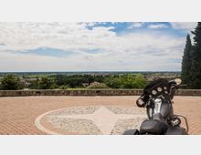 Terrazza panoramica pavimentata in mattoni con una rosa dei venti in pietra al centro e muretto di parapetto affacciato su una vasta pianura verde con case sparse. Sullo sfondo cielo parzialmente nuvoloso; a destra si vede una motocicletta parcheggiata e alberi alti.