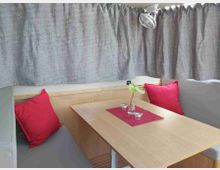 Angolo pranzo interno con tavolo in legno tra divanetti imbottiti grigi e cuscini rossi. Tende grigie chiuse lungo le pareti e lampada orientabile sopra il tavolo, con runner rosso, piccolo vaso con fiore e bicchierini.