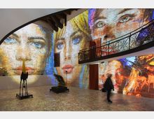 Interno di una galleria o museo con ampie pareti in mattoni su cui sono proiettati grandi ritratti colorati che coprono quasi tutta la superficie. Lo spazio ha un soppalco con ringhiera in ferro battuto, tende rosse e alcune sculture esposte su piedistalli sul pavimento in pietra.