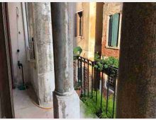 Loggiato con grandi colonne in pietra e pavimento in marmo, affacciato su un cortile interno tramite una ringhiera in ferro battuto. Sullo sfondo si vedono facciate di edifici in intonaco e mattoni con finestre e vegetazione nel cortile.
