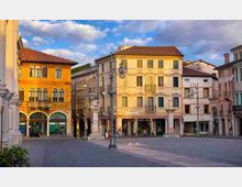 Piazza urbana con edifici storici a più piani dalle facciate color ocra e gialla, con portici ad archi, balconi e finestre allineate. Lampioni in ferro e pavimentazione in pietra delimitano lo spazio aperto sotto un cielo parzialmente nuvoloso.