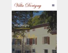 Facciata di una villa di campagna a due piani con intonaco chiaro e persiane in legno, con finestre rettangolari e tetto a falde. L’edificio è parzialmente incorniciato da rami e alberi, con vegetazione sullo sfondo.
