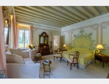 Camera da letto in stile classico con soffitto a travi dipinte, pareti affrescate e grande letto con testiera decorata. Arredi d’epoca, poltrone e divano chiaro, comodini con lampade e ampia finestra con tende che affaccia sul giardino.