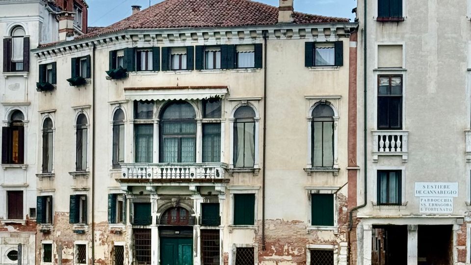 Facciata di edifici storici affacciati su un canale, con intonaco chiaro parzialmente scrostato, tetti in coppi rossi e persiane scure. Al centro si vede un portale ad arco con balcone e finestre ad arco, mentre in primo piano scorre l’acqua del canale.