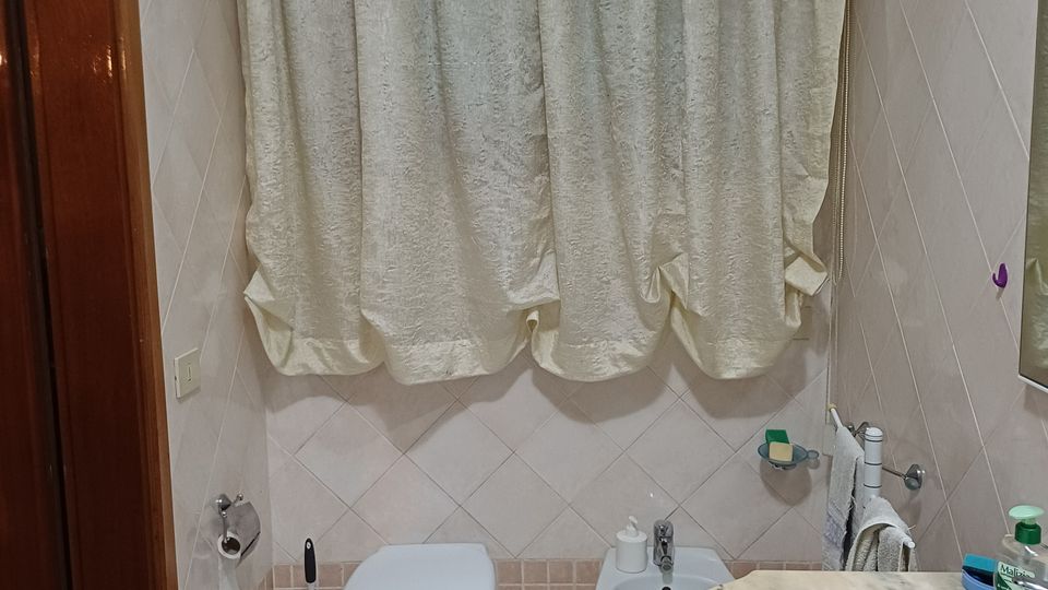 Bagno domestico con pareti e pavimento piastrellati in tonalità beige, wc sospeso e bidet affiancati sotto una grande finestra coperta da tende chiare. In primo piano si vede un lavabo incassato in un piano in marmo, con porta in legno sul lato sinistro.