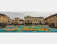 Piazza storica con ampio tappeto floreale multicolore (infiorata) al centro e aiuole lungo i bordi. Sullo sfondo si affacciano edifici in stile italiano con portici ad archi, facciata centrale con orologio e balconi, sotto un cielo nuvoloso.