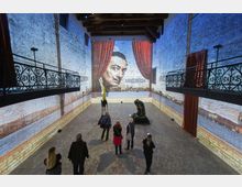 Ampia sala espositiva in un edificio industriale riconvertito, con pareti in mattoni a vista e due balconate laterali in ferro battuto sotto un soffitto con travi in legno. Sul fondo una grande proiezione murale con un volto incorniciato da tende rosse, con sculture esposte al centro dello spazio.