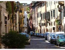 Strada stretta in acciottolato in un centro storico, fiancheggiata da edifici a più piani con persiane e arcate al piano terra. Auto parcheggiate su entrambi i lati, lampioni e facciate con balconi e vegetazione lungo la via.
