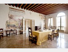 Sala da pranzo storica con soffitto a travi in legno a vista, pareti con affreschi e pavimento in terrazzo. Al centro un lungo tavolo con tovaglia gialla e sedie in legno, illuminato da una grande finestra ad arco con tende chiare.