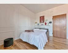 Camera da letto moderna e luminosa con pavimento in legno, letto matrimoniale con copriletto bianco e pareti bianche con boiserie. Sopra il termosifone c’è una mensola con libri e decorazioni, un poster a parete e una porta in legno sulla destra.