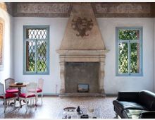 Salone interno con grande camino in pietra al centro e fregi decorativi sul soffitto. Due finestre con infissi azzurri e griglie a rombi affacciano sul verde; pavimento in seminato veneziano con tavolino e sedie a sinistra e divano in pelle a destra.