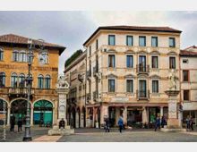 Piazza urbana con edifici storici a più piani dai colori ocra e crema, con finestre ornate, portici ad archi al piano terra e tetti in coppi. In primo piano si vedono due colonne monumentali con statue e un lampione in ferro battuto, con caffè e negozi lungo i lati della piazza.