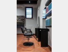 Interno di un salone di parrucchiere con due poltrone nere da taglio, postazioni con specchio e scaffali a parete pieni di prodotti per capelli. Pavimento in cotto, pareti bianche e finestre alte con griglie metalliche, con lampade a soffitto e climatizzatore.