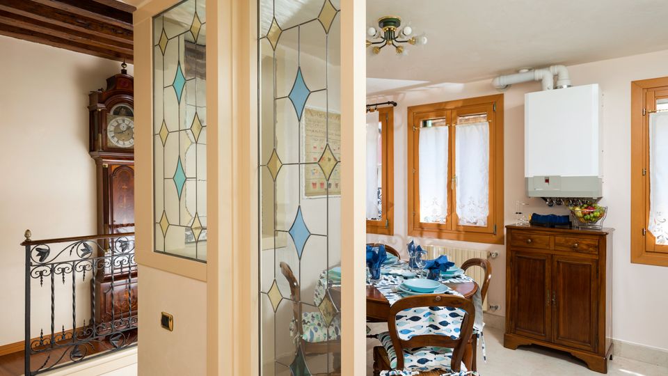Interno domestico con zona pranzo e cucina: tavolo apparecchiato accanto a finestre con infissi in legno e tenda chiara, e mobile credenza sotto una caldaia a parete. In primo piano una parete con vetri decorati a motivi geometrici e, sul lato, travi in legno e ringhiera in ferro battuto con un orologio a pendolo.
