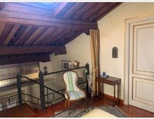 Soppalco interno con soffitto inclinato e travi in legno a vista, affacciato su una scala con ringhiera in ferro e parete rivestita di piastrelle decorative. Sul pavimento in legno ci sono una poltrona a righe, un piccolo tavolino e una tenda accanto a una porta con cornici.