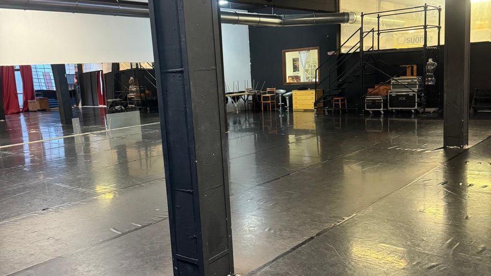 Ampio spazio interno tipo sala prove o studio teatrale con pavimento nero lucido e grandi colonne metalliche. Dal soffitto scuro pendono fari e tubazioni, con un soppalco laterale e alcune attrezzature e sedie lungo le pareti.