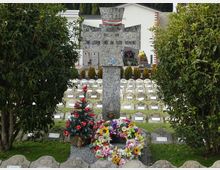 Monumento commemorativo in pietra a forma di croce in un cimitero militare, con corona di fiori e addobbi alla base. Sullo sfondo si vedono file ordinate di piccole lapidi e un edificio chiaro, incorniciati da siepi e alberi sempreverdi.
