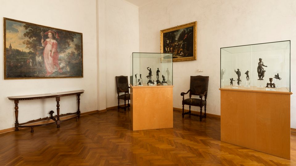 Sala museale con pareti bianche e pavimento in parquet a spina di pesce, con due vetrine su piedistalli in legno che espongono piccole sculture. Alle pareti sono appesi grandi dipinti incorniciati e lungo i lati si vedono un tavolo consolle e alcune sedie d’epoca.