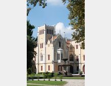 Facciata di una villa in stile neogotico con torre merlata, finestre ad arco e tetti in tegole, affacciata su un giardino con prato e vialetti. In primo piano si vede una fontana in pietra, con alberi ai lati che incorniciano l’edificio.