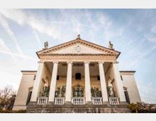 Facciata di una chiesa in stile neoclassico con ampio portico a sei colonne e frontone triangolare con stemma e statue sul tetto. L’edificio chiaro è sopraelevato su una scalinata in pietra, con balaustra e vegetazione rampicante, sullo sfondo di un cielo azzurro.