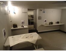 Interno di un bunker o rifugio sotterraneo con pareti e soffitto in cemento, illuminazione essenziale e tavoli con mappe e strumenti. Sul fondo una porta aperta conduce a una stanza con letti a castello, mentre mensole laterali espongono oggetti e poster informativi.