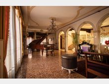 Ampio salone interno in stile classico con pavimento in terrazzo, grandi finestre con tende a sinistra e lampadario centrale. Nella stanza si vedono un pianoforte a coda, poltrone e sedute, e una serie di archi che conducono a un’area con scala sullo sfondo.