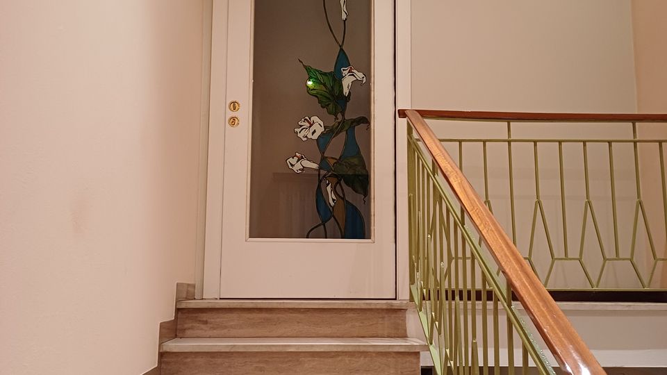Vano scale interno con gradini in marmo chiaro e alzate effetto legno, affiancato da una ringhiera verde con corrimano in legno. In cima alle scale c’è una porta bianca con pannello in vetro decorato a motivo floreale e una finestra orizzontale sopra la porta.