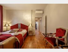 Camera da letto in stile classico con letto matrimoniale, testiera imbottita rossa e comodini con lampade ai lati, su pavimento in legno. A destra ci sono una poltrona rossa e un portavaligie, mentre in fondo si vede una porta aperta verso un corridoio con climatizzatore sopra.