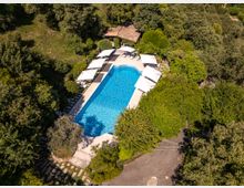 Vista aerea di una piscina all’aperto a forma rettangolare con estremità arrotondate, circondata da una terrazza con ombrelloni bianchi e lettini. L’area è immersa nel verde di alberi e siepi, con un piccolo edificio dal tetto in tegole vicino al bordo della piscina.