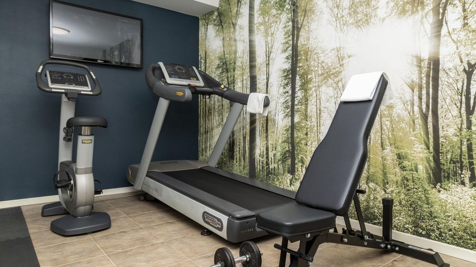 Piccola sala palestra con cyclette e tapis roulant davanti a una parete blu con TV a schermo piatto. Sulla parete laterale c’è una grande carta da parati con bosco illuminato dal sole, e in primo piano una panca regolabile con manubri su pavimento piastrellato.
