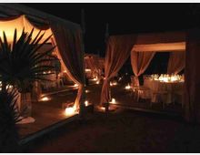 Area lounge all’aperto di notte con gazebo in tessuto chiaro e tende drappeggiate su pedane in legno. Tavoli apparecchiati e sedie bianche sono illuminati da numerose candele disposte lungo i percorsi.