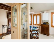 Interno domestico con zona pranzo e cucina: tavolo apparecchiato accanto a finestre con infissi in legno e tenda chiara, e mobile credenza sotto una caldaia a parete. In primo piano una parete con vetri decorati a motivi geometrici e, sul lato, travi in legno e ringhiera in ferro battuto con un orologio a pendolo.