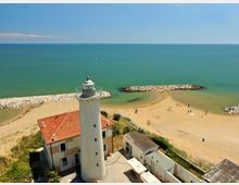Veduta aerea di un faro bianco con lanterna e ringhiera, affiancato da edifici con tetto in tegole rosse, su una spiaggia sabbiosa. Sullo sfondo il mare calmo e due frangiflutti in pietra che proteggono una piccola insenatura.