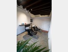 Interno di un salone da parrucchiere con due postazioni lavatesta e sedie nere, pavimento in piastrelle grigie e pareti bianche. Soffitto con travi in legno a vista e mobile lungo la parete con prodotti per capelli.