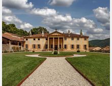 Villa storica a due piani con facciata simmetrica, portico centrale con colonne e tetto in tegole rosse, circondata da un giardino curato. In primo piano un vialetto di ghiaia si dirama tra prati verdi, con colline boscose e cielo nuvoloso sullo sfondo.