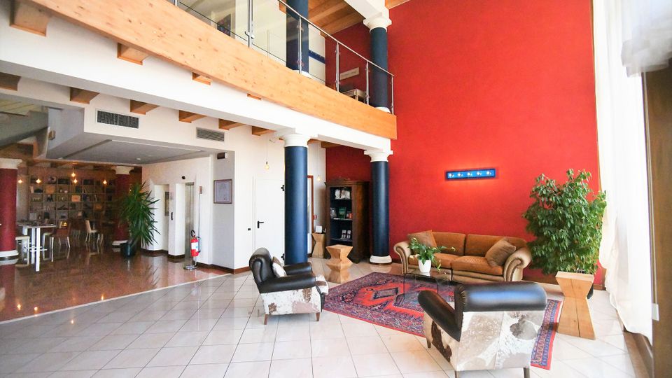 Ampia hall interna con soffitto alto e soppalco in legno con parapetto in vetro, parete rossa e colonne scure. Area salotto con divano, poltrone, tappeto e piante su pavimento piastrellato, con zona bar/sala sullo sfondo.