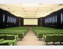 Interno di una sala cinema con molte file di poltrone verdi disposte a ventaglio verso un grande schermo bianco. Pareti scure con pannelli luminosi verticali e soffitto chiaro con illuminazione incassata.