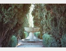 Viale di giardino con sentiero in ghiaia fiancheggiato da alte siepi e cipressi, che forma un corridoio verde. Al centro si trova una fontana in pietra con ampia vasca circolare.