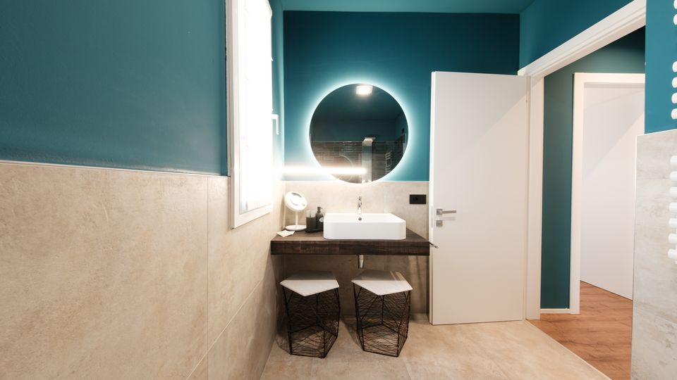 Bagno moderno con pareti verde petrolio e rivestimento in piastrelle beige, lavabo rettangolare su piano in legno scuro e grande specchio rotondo retroilluminato. Accanto a una finestra luminosa ci sono due sgabelli a rete sotto il piano e una porta bianca aperta verso un corridoio con pavimento in legno.