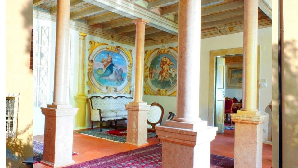 Portico interno di una villa storica con colonne in pietra e soffitto a travi dipinte, pavimento in cotto e tappeti. Sul fondo si vedono pareti affrescate con grandi medaglioni decorativi e un passaggio verso una sala arredata.
