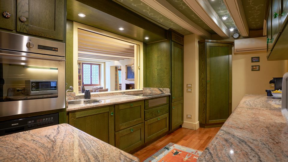 Cucina interna con mobili in legno scuro e piani di lavoro in granito, dotata di forno incassato, lavello e illuminazione a faretti. Un’apertura collega alla zona giorno con travi a vista e finestra a riquadri, mentre il pavimento è in legno con un tappeto al centro.