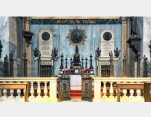 Altare interno di una chiesa con parete in marmo azzurro e iscrizione “DEO UNI AC TRINO” in alto, affiancata da due grandi lapidi commemorative. Al centro si vedono candelabri e decorazioni metalliche, una cancellata in ferro battuto e un piccolo altare con tovaglia bianca e gradini rossi, con balaustre e panche in primo piano.