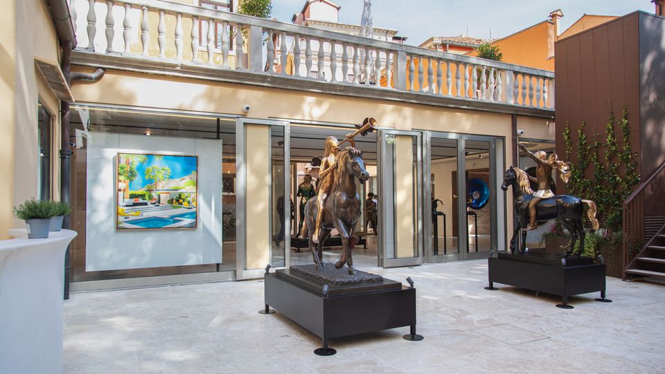 Cortile di una galleria d’arte con pavimentazione in pietra chiara e grandi porte a vetri aperte su sale espositive illuminate. All’esterno sono esposte due sculture equestri su basamenti e un dipinto appeso alla parete, con balconata in pietra al piano superiore e edifici circostanti.