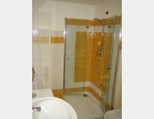 Bagno con box doccia ad angolo in vetro e profili metallici, piatto doccia bianco e soffione con doccetta. Pareti rivestite di piastrelle bianche con fasce arancioni e termosifone a scaletta sulla sinistra.
