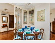 Sala da pranzo in un interno domestico con tavolo rotondo in legno apparecchiato, tovaglia a motivi blu e sedie coordinate su pavimento in parquet. Sullo sfondo si vedono pannelli vetrati decorati con motivi geometrici, una cornice con specchio, un piccolo tavolino con fiori e un quadro a parete.