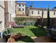 Piccolo giardino privato con prato curato, alcune piante in vaso e fioriere lungo il muro, e una sedia a sdraio appoggiata vicino alla recinzione. Sullo sfondo si vedono edifici residenziali a più piani e un camminamento in legno con tavolo e sedie sul lato destro.